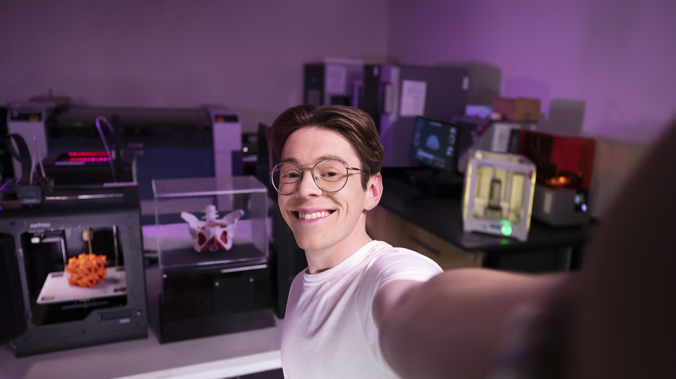 Male student smiling at the camera taking a selfie style photo, in the background shows a dimly lit science lab.