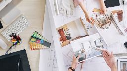 Two sets of hands compare design documents over a desk crowded with design utensils including a ruler, colour swatch, computer and tablet.