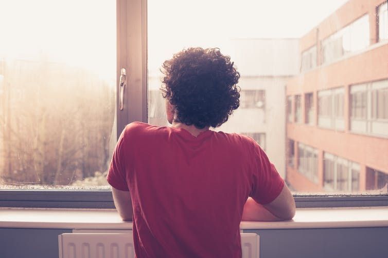 STOCK Lonely in lockdown man at window Man in a red T-shirt looking out of the window during COVID-19 lockdown