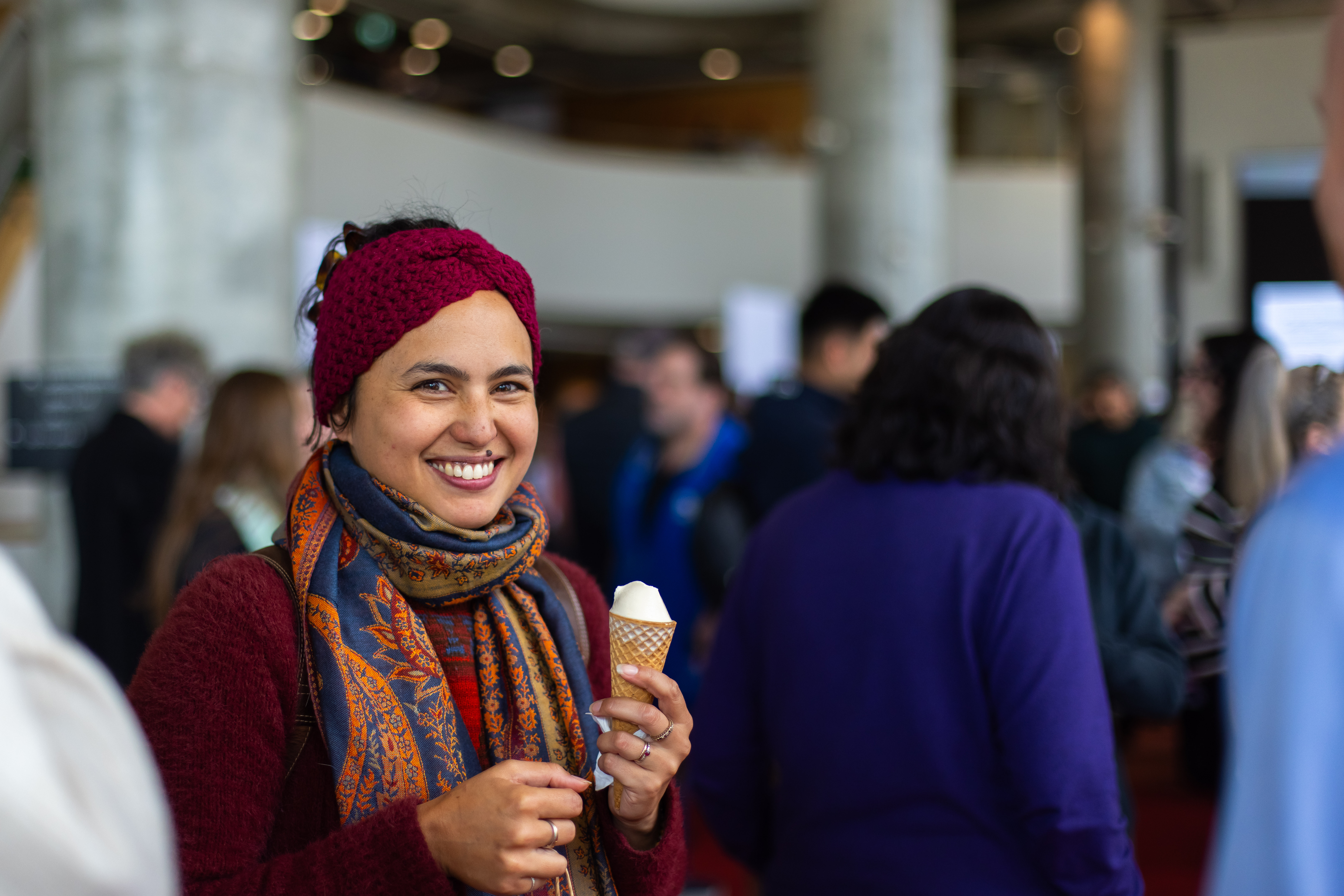 Woman smiling with an ice-cream in a busy indoor event space, wearing a red headband, patterned scarf and maroon sweater.
