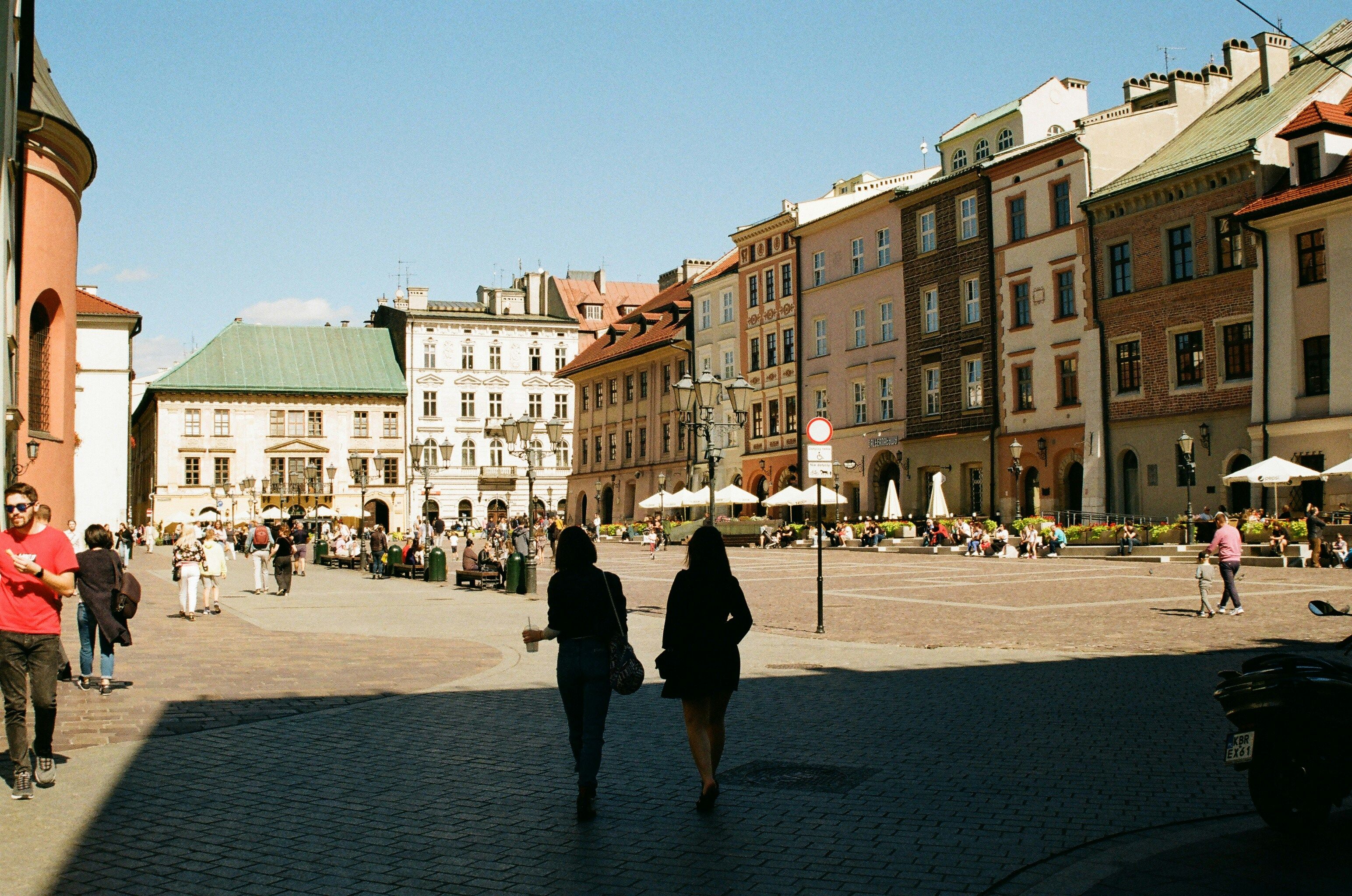 Main Market Square Krakow Poland A shadow of two people enjoying a drink walking through an old European town square surrounded by old buildings.