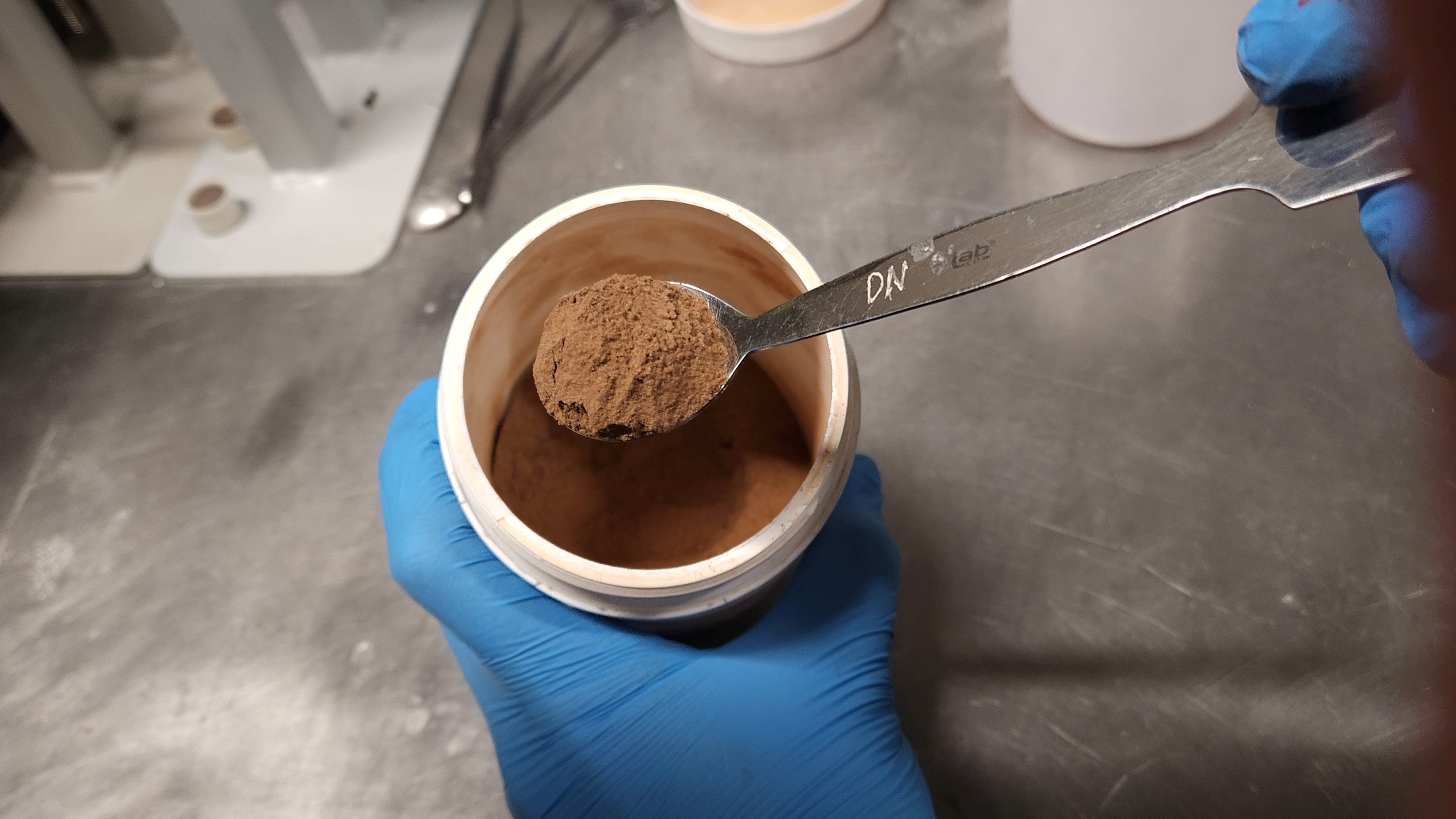 Martian regolith simulant, made to mimic the surface composition at Gale Crater on Mars. A spoonful of fine, red dust is held above a container holding the material. The dust is regolith simulant, made to mimic the surface composition found at Gale Crater on Mars.