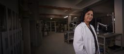 Landscape shot of woman in science laboratory wearing a lab coat, science equipment is seen in the background. 