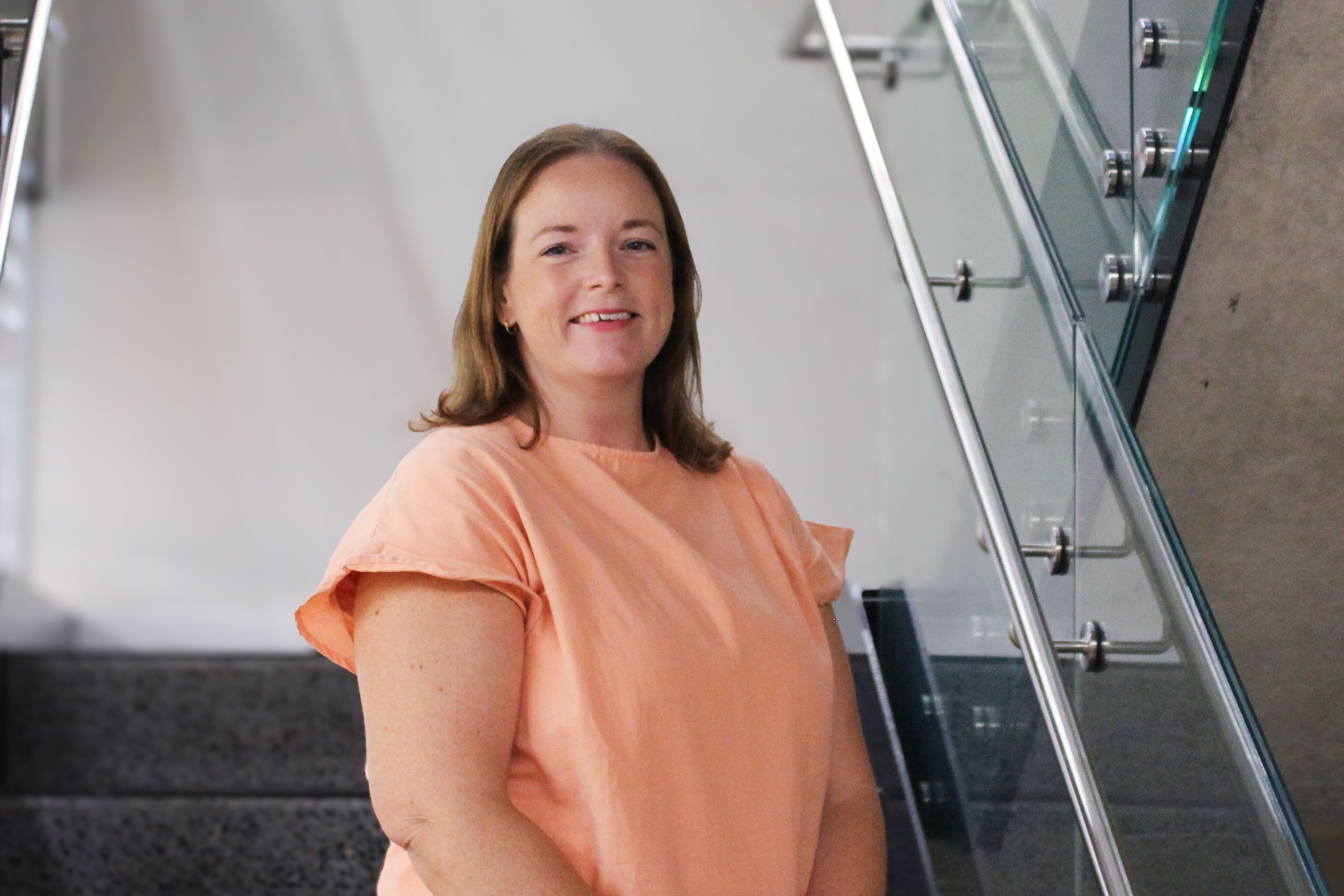 Swinburne Staff Member Sarah Jellie standing on a set of concrete stairs with a glass railing and a white wall behind. 