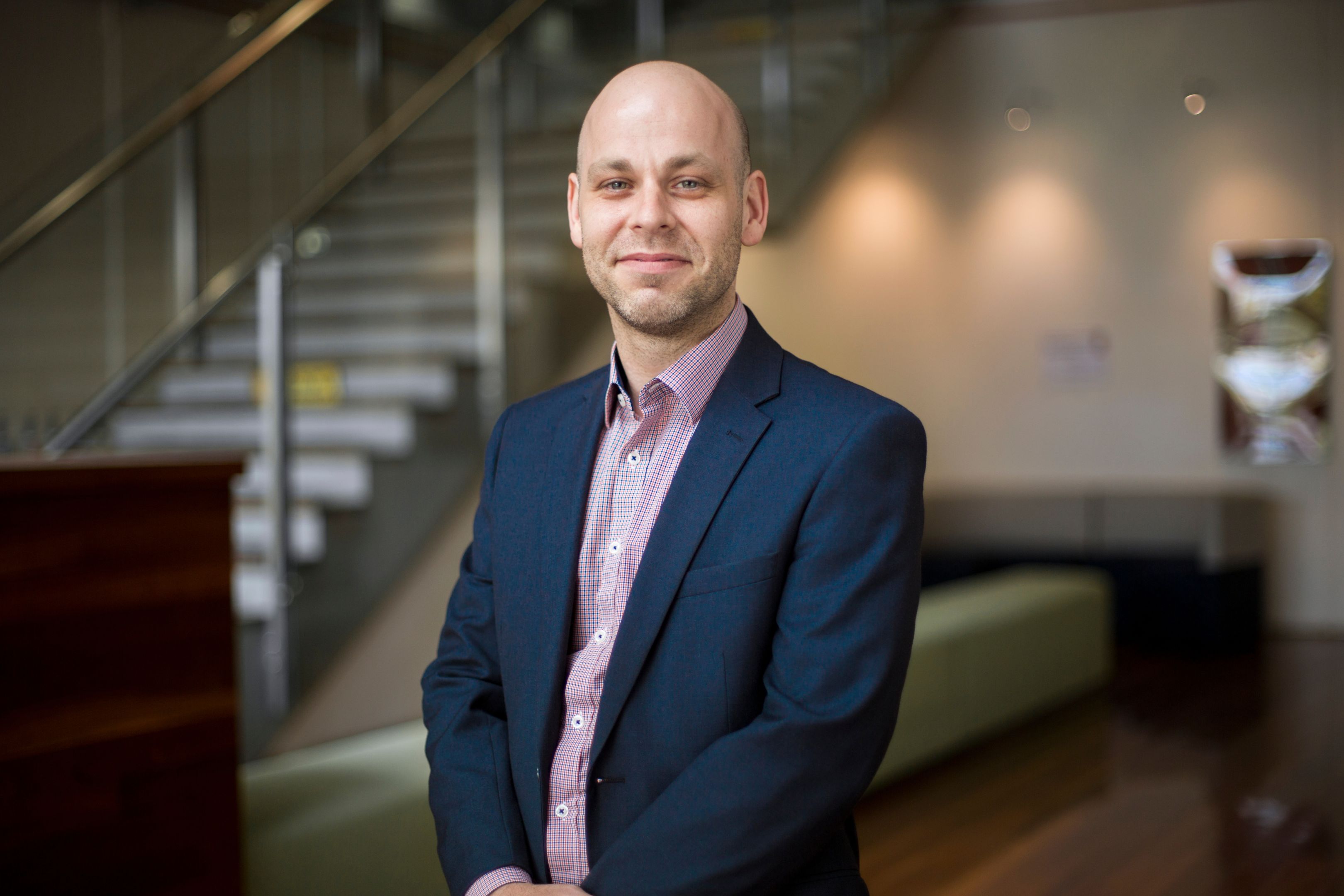 Travis Keating Travis Keating wearing suit attire poses with hands clasped in front in the foyer of the SPS building