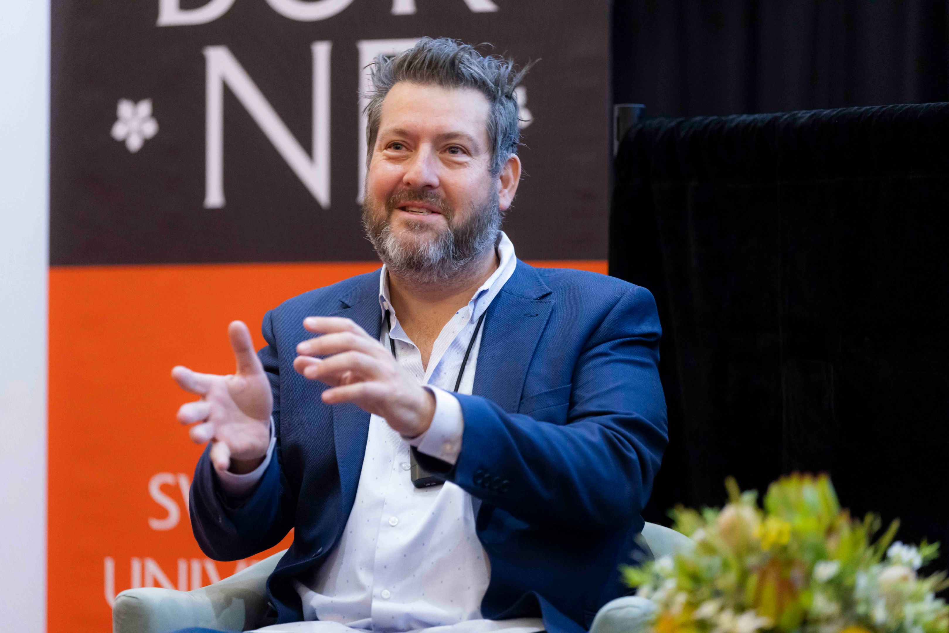 Jefferson Harcourt delivers AGSE Oration 2022 Man in blue blazer and white shirt sits in armchair in front of Swinburne banner as he delivers AGSE Oration