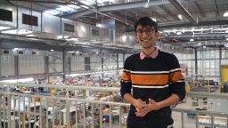  A tall man in a black long jumper with a horizontal orange stripe is pictured in a factory environment with machines in the background