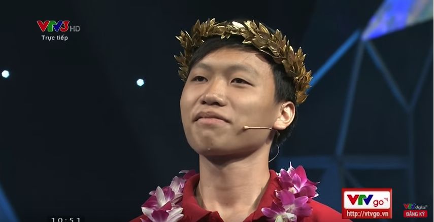 Road to Olympia Peak winner 2017 High school student wearing crown of gold leaves, flower chain around his neck and microphone/earpiece. TV logos and time stamp watermarked on shot.