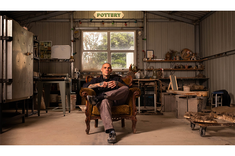 Person seated in an armchair inside a pottery studio with shelves, tools and ceramic materials.