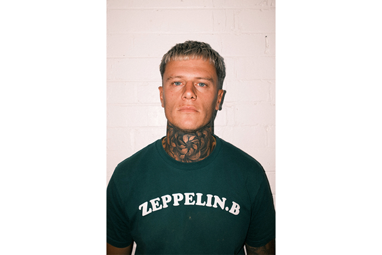 Portrait of a person wearing a black T-shirt standing against a pale brick wall.