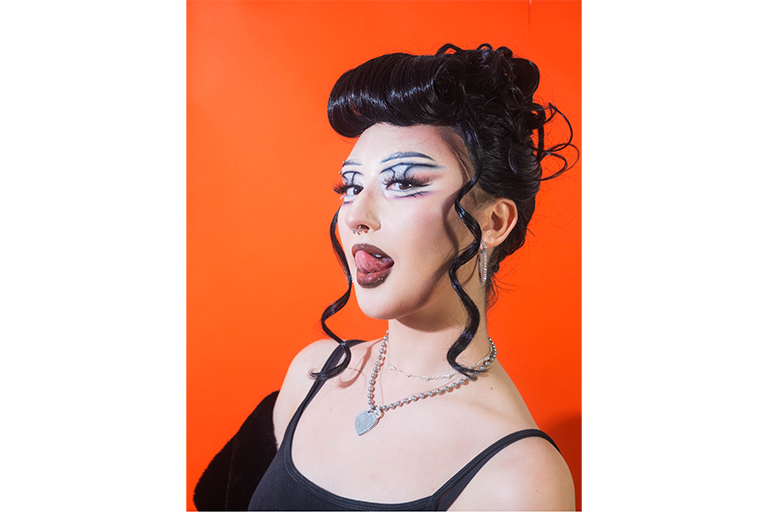 Portrait of a person with bold makeup, dark curled hair and jewellery against an orange background.
