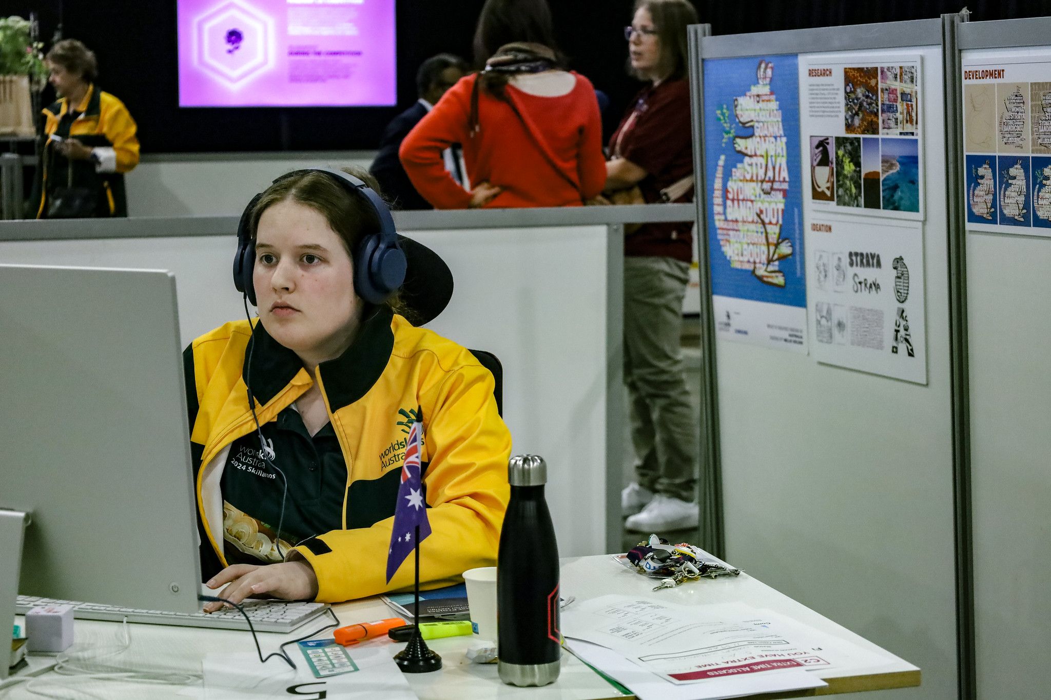 Nellie Holder competing in Graphic Design WorldSkills Nellie Holder sitting at a computer. She is wearing a yellow "Skillaroos" jacket and headphones. She is concentrating intently.