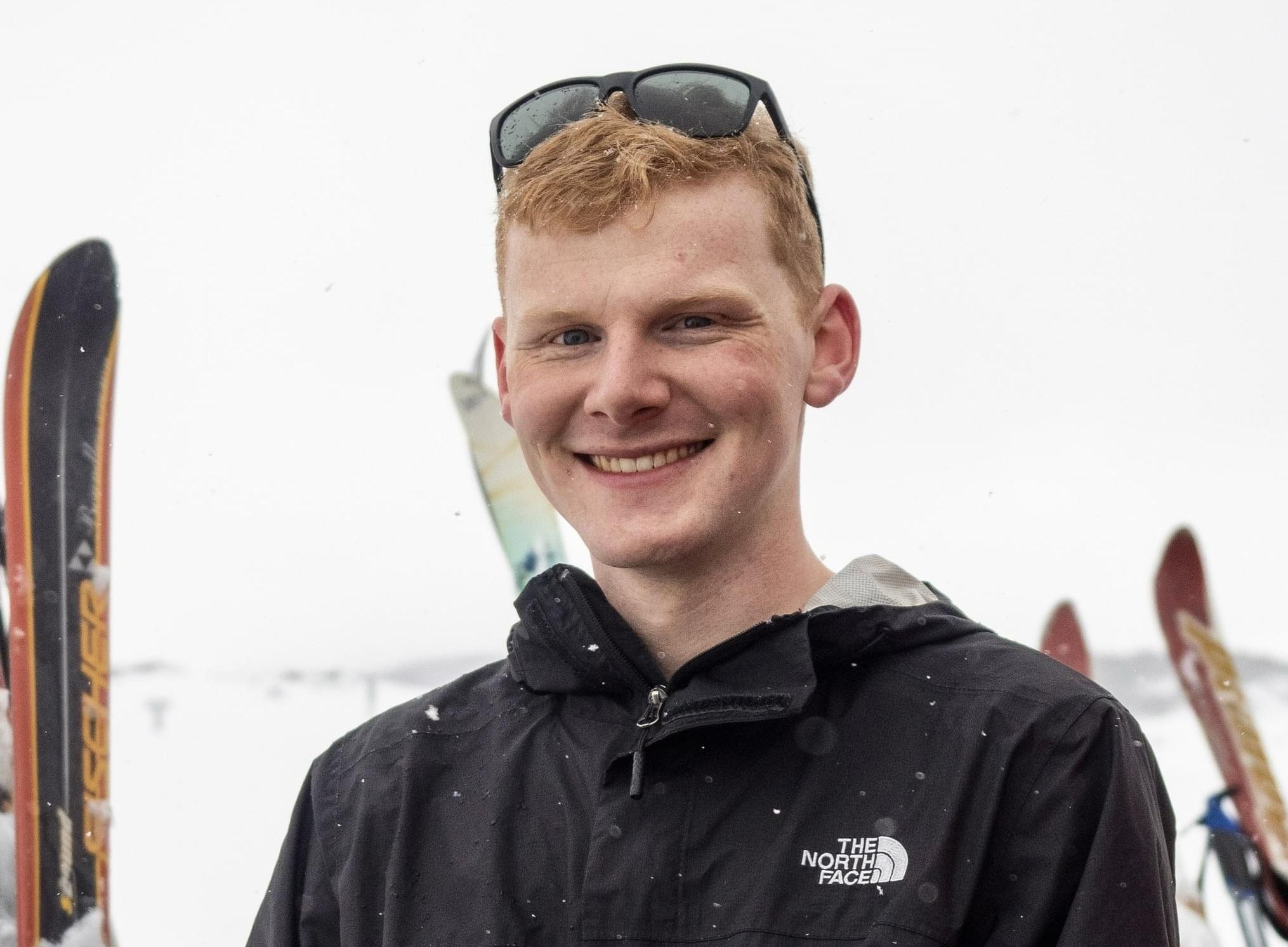 Placement student Nic A red-haired student in a black snow jacket, smiling against a backdrop of skis and snow.