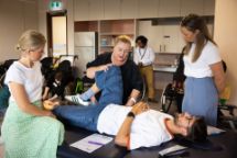 A student lying on a table being shown by his teacher how to assist someone stretch in a classroom with other students.