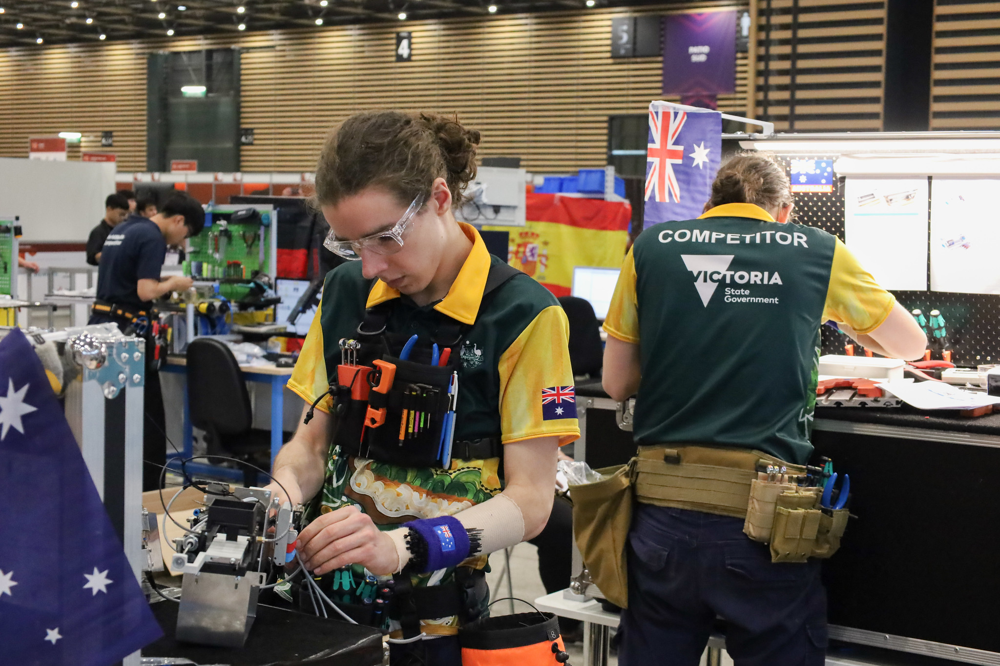 Will is facing the camera and working on a piece of electronics in front of him. He is wearing protective goggles. Behind him Magnus is bent over a table covered in tools. They are both wearing "skillaroos" polo shirts and tool belts. They are deep in concentration.