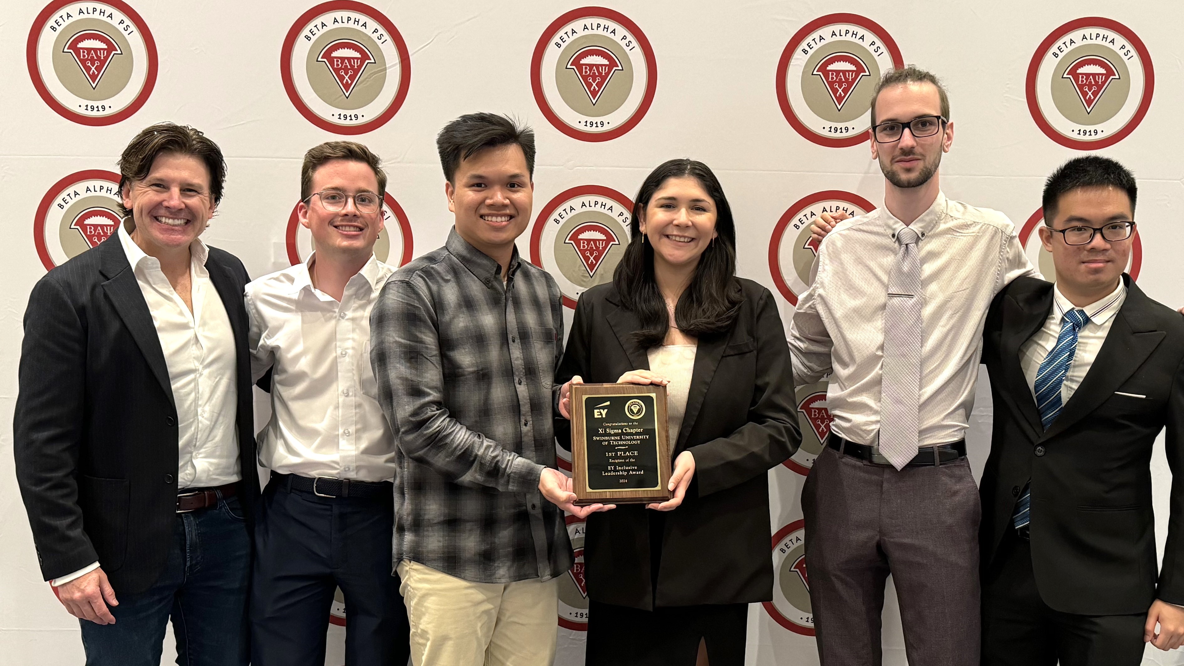 Five students and an academic stand in front of a photo wall holding a plaque