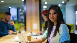 Female student is seen studying in a communal student study space, other students in background.